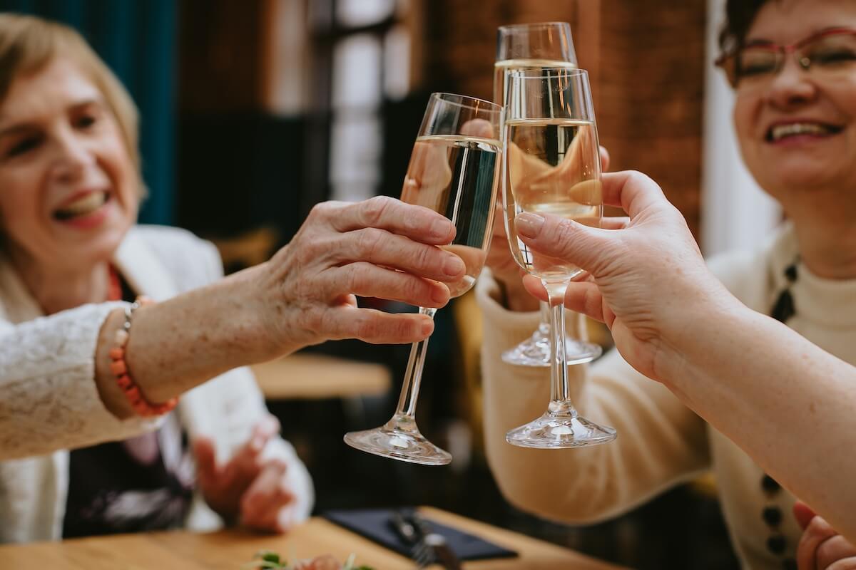 Hands of smiling elderly women clinking glasses of wine or champagne while celebrating their meeting or some holiday in cozy cafe or restaurant