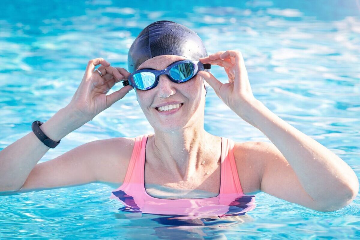 jpeg-optimizer_shutterstock_2394531853 A smiling older woman adjusts her swim goggles in a bright blue pool, enjoying the kind of active aquatic lifestyle that water aerobics for seniors at Lindell Living makes possible every day.