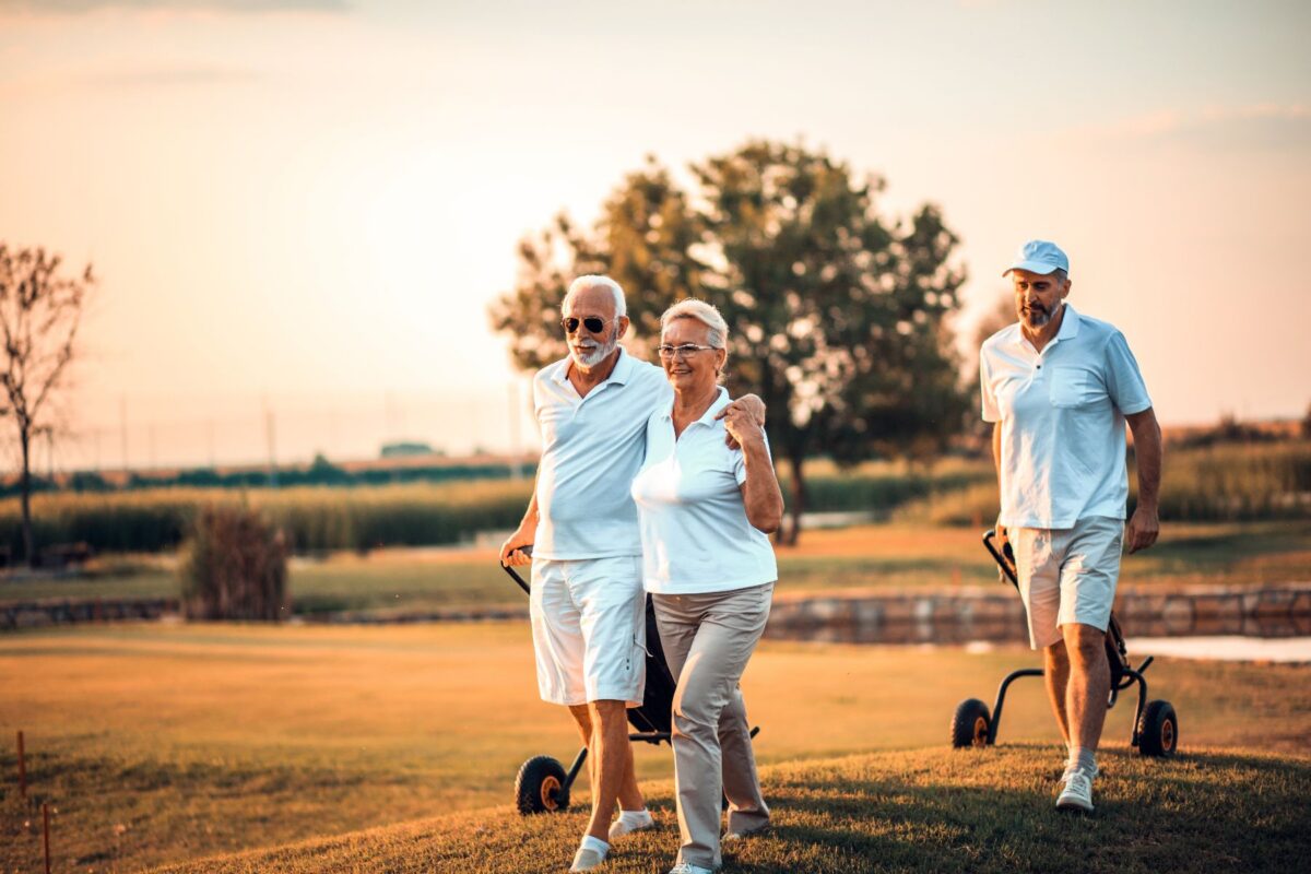 Making friends after 50: three older adults walk together on a golf course at sunset, laughing and chatting.